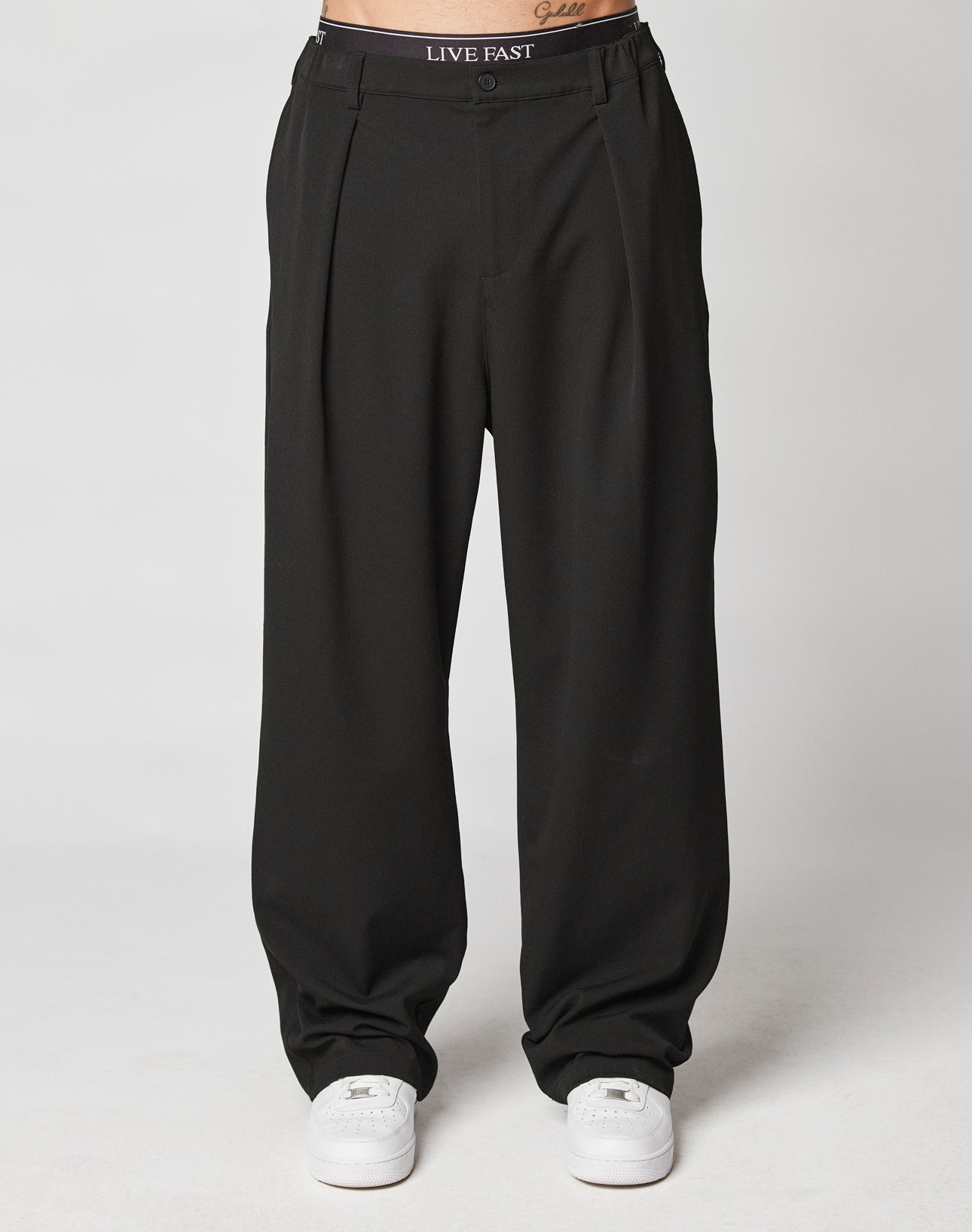 Someone stands against a plain white background wearing LIVE FAST DIE YOUNG Wide Casual Pants—loose black pleated trousers with a LIVE FAST waistband—paired with white sneakers.| model