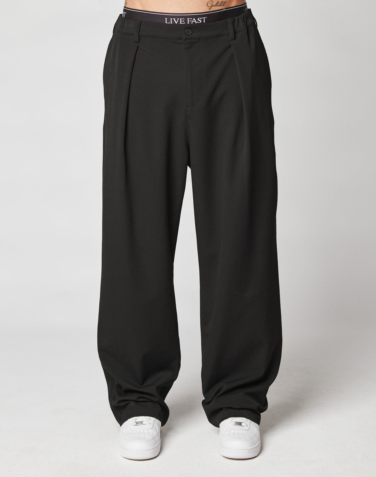 A person wears LIVE FAST DIE YOUNG Wide Casual Pants in black with side pockets and an elastic waistband that reads LIVE FAST. They pair the look with white sneakers against a plain white background.