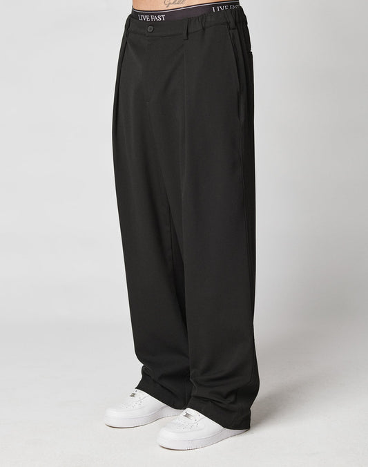 A person wears loose-fitting black Wide Casual Pants by LIVE FAST DIE YOUNG, featuring a printed waistband, paired with white sneakers. Only the lower body is shown against a plain light background.