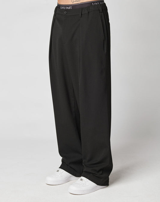 A person wears LIVE FAST DIE YOUNG Wide Casual Pants—loose, wide-fit black trousers with side pockets and an elastic waistband featuring LIVE FAST—paired with white sneakers; only the lower half is shown against a light background.