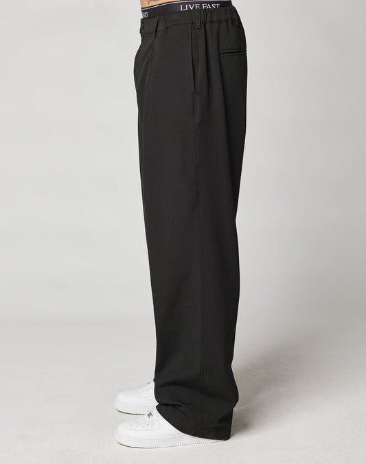 A person stands sideways against a plain light background, wearing LIVE FAST DIE YOUNG Wide Casual Pants, white sneakers, and a visible black waistband with "LIVE FAST" text.
