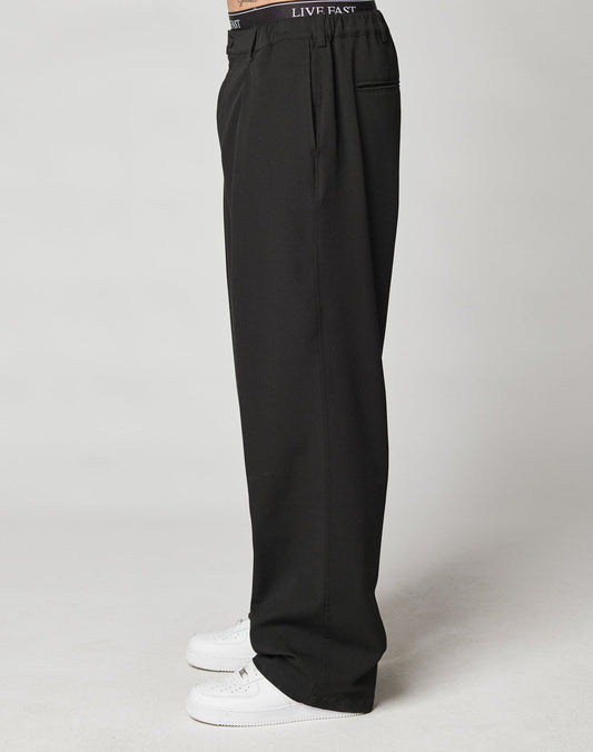 A person stands in profile wearing LIVE FAST DIE YOUNG Wide Casual Pants with an elastic waistband and LIVE FAST visible, paired with white sneakers against a plain light background.
