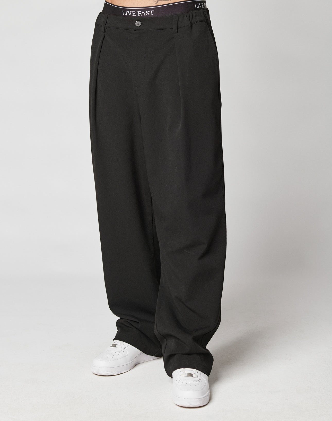 A person wearing loose-fitting Wide Casual Pants by LIVE FAST DIE YOUNG with wide fit, elastic waistband, side pockets, and labeled LIVE FAST, paired with white sneakers, stands against a plain light background. Upper body not visible.