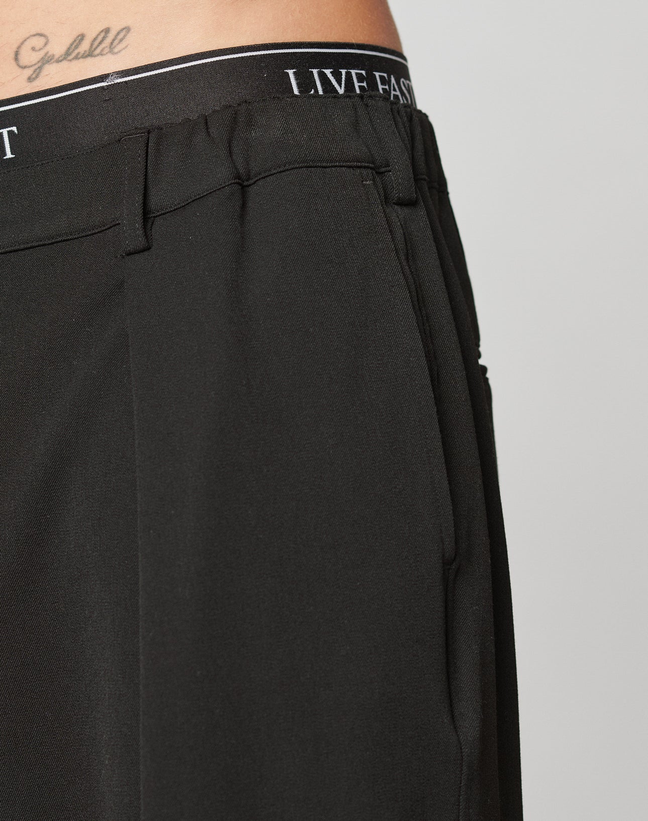 Close-up of a person wearing Wide Casual Pants by LIVE FAST DIE YOUNG, featuring a black wide-fit design with an elastic waistband and LIVE FAST in white. Part of a Gabrielle tattoo is visible on their lower abdomen.