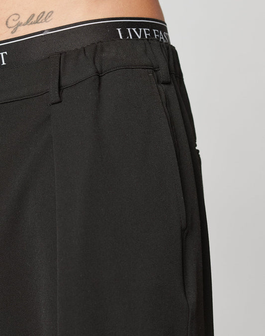 Close-up of a person wearing black Wide Casual Pants by LIVE FAST DIE YOUNG, featuring LIVE FAST on the waistband and a cursive Goodluck tattoo above the waist. Background is plain and light-colored.