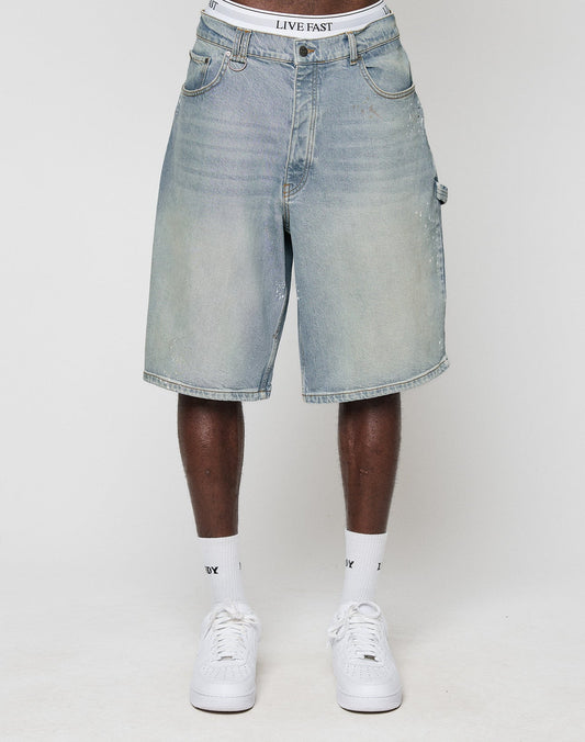 A person wears LIVE FAST DIE YOUNG Carpenter Jorts in a baggy fit, light blue, knee-length style with visible LIVE FAST waistband, paired with white sneakers and socks, standing against a plain white background.