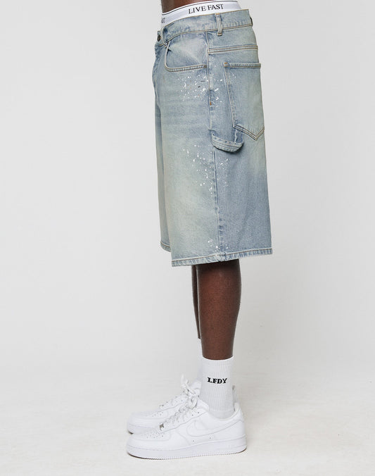 A person stands sideways against a plain background wearing LIVE FAST DIE YOUNG Carpenter Jorts in light blue, featuring Farbspritzer detail and Baggy Fit, paired with a visible LIVE FAST waistband, white LFDY socks, and Nike Air Force 1 sneakers.