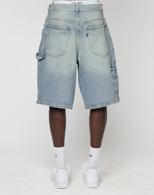 A person is seen from behind wearing LIVE FAST DIE YOUNG Carpenter Jorts in light blue baggy fit denim with large back pockets, styled with white socks and sneakers, set against a plain background.