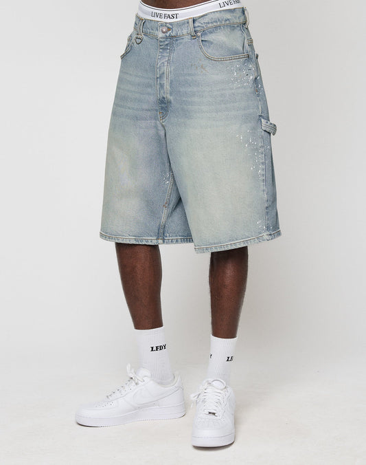 A person wearing LIVE FAST DIE YOUNG Carpenter Jorts in light blue 100% cotton with LIVE FAST on the waistband, white LFDY socks, and white sneakers stands against a plain white background.