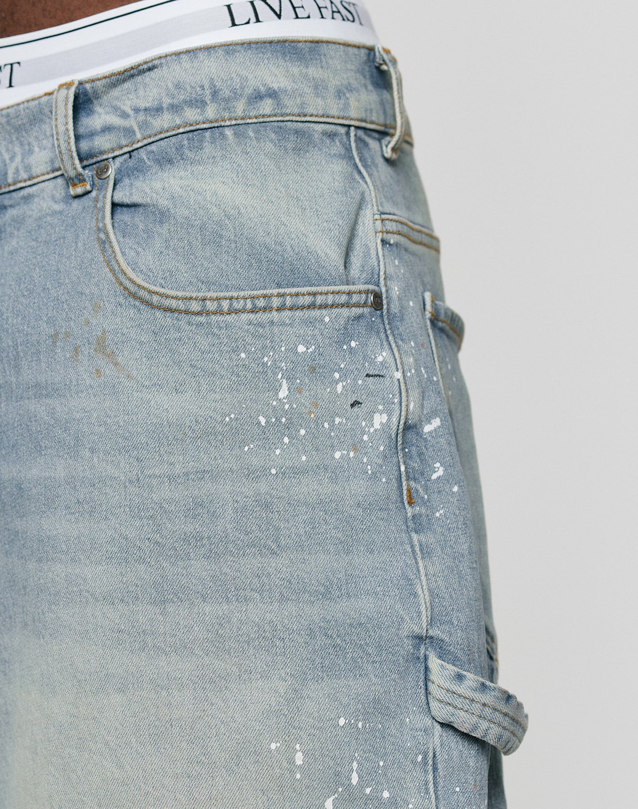 Close-up of LIVE FAST DIE YOUNG Carpenter Jorts in baggy light blue denim with white paint splatters and a utility pocket. The white LIVE FAST waistband of underwear is visible above these 100% cotton shorts.