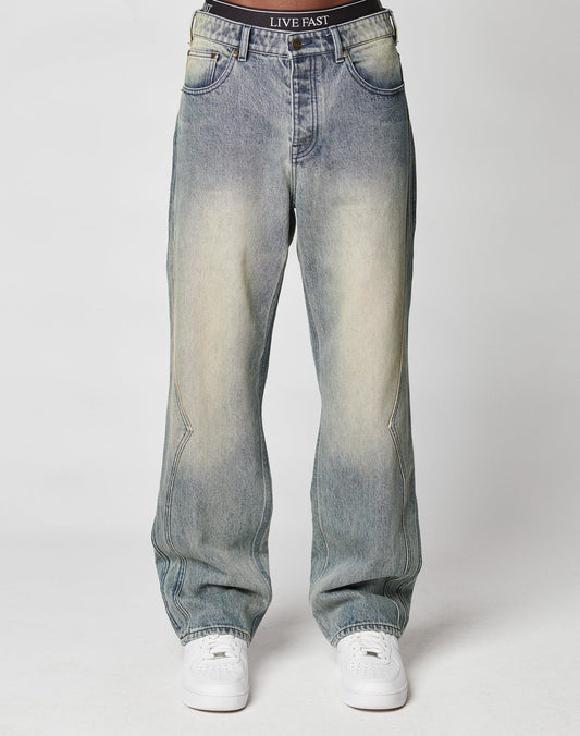 A person wears the LIVE FAST DIE YOUNG Rhomb Wide Denim—faded blue, high-waist, 100% Baumwolle jeans with diamond panels and visible front pockets—in a baggy fit, styled with sneakers against a plain white background. Only lower torso and legs shown.