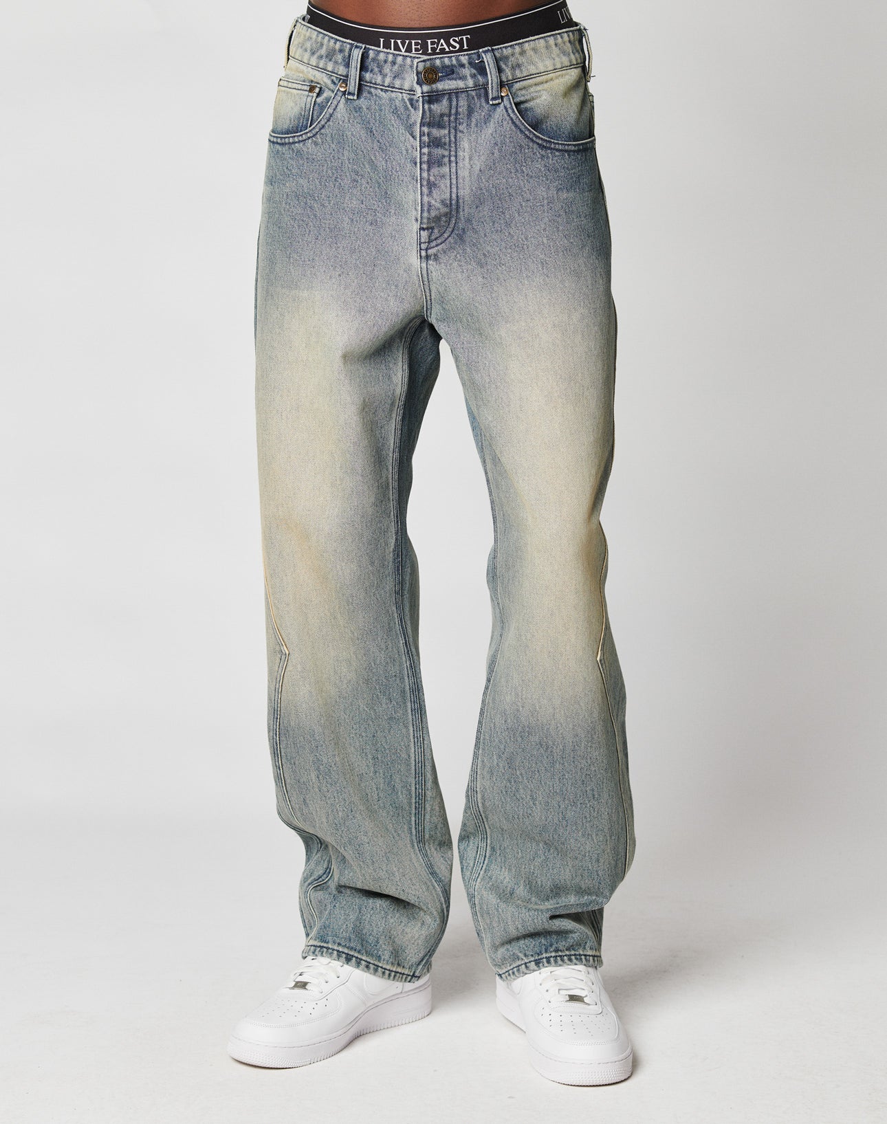Someone wears LIVE FAST DIE YOUNGs Rhomb Wide Denim—baggy, light blue faded jeans with LIVE FAST on the waistband—paired with white sneakers, standing against a plain white background.