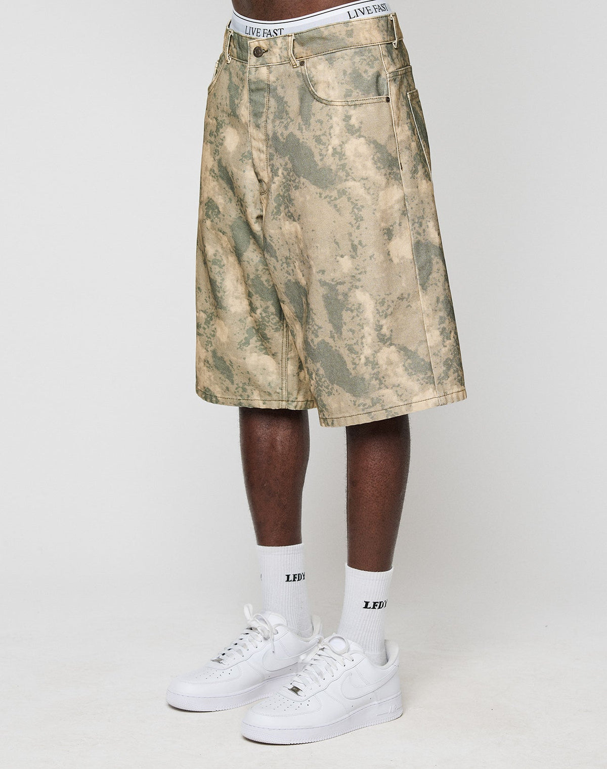 A person in LIVE FAST DIE YOUNG Canvas Shorts with a baggy fit and straight leg, white socks with LEDF text, and white Nike Air Force 1s stands against a plain white background. | model