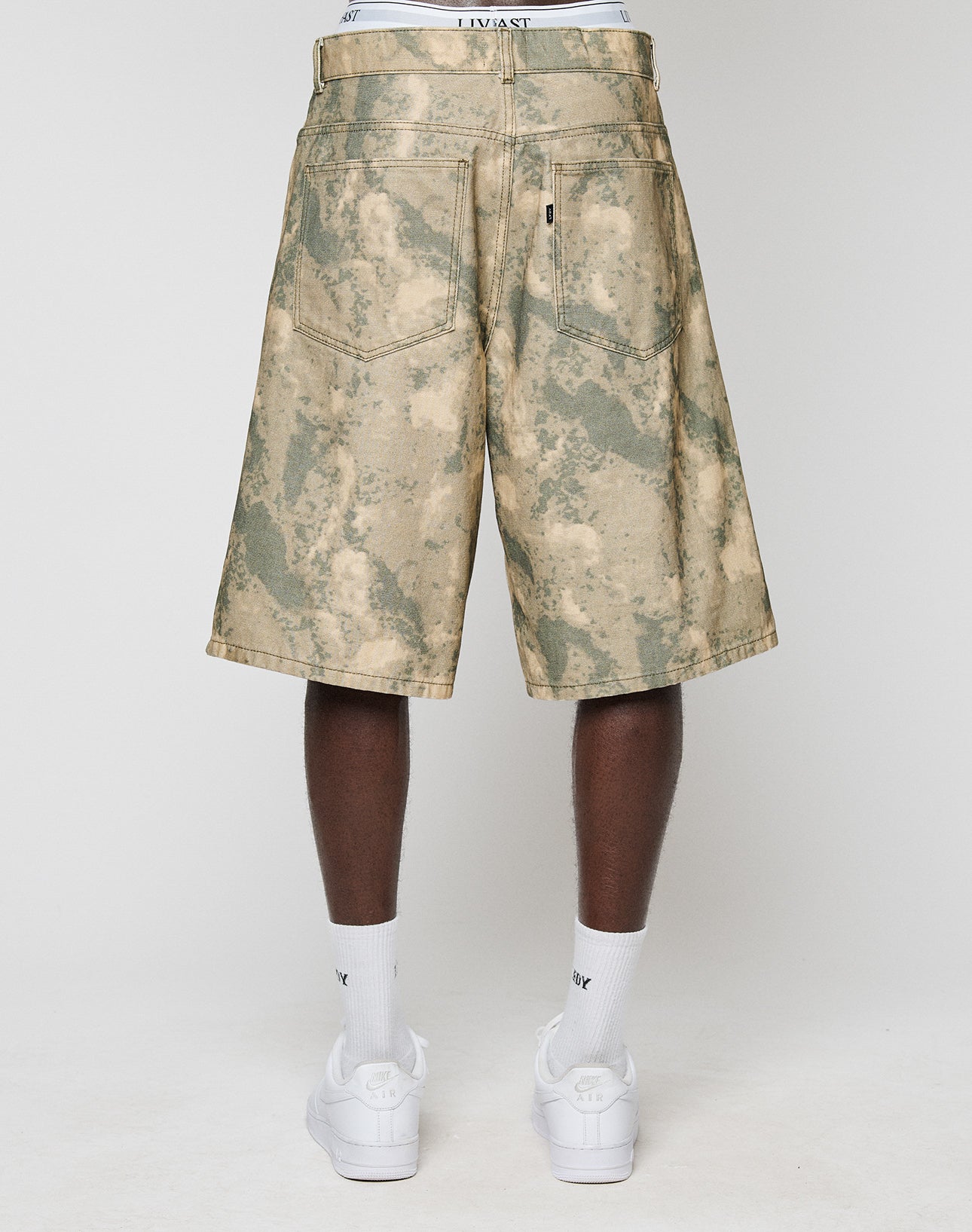 A person faces away, wearing LIVE FAST DIE YOUNG Canvas Shorts (100% Baumwolle, Baggy Fit, gerade Bein) with back pockets. The white waistband of underwear shows above the shorts, paired with white Nike socks and sneakers.