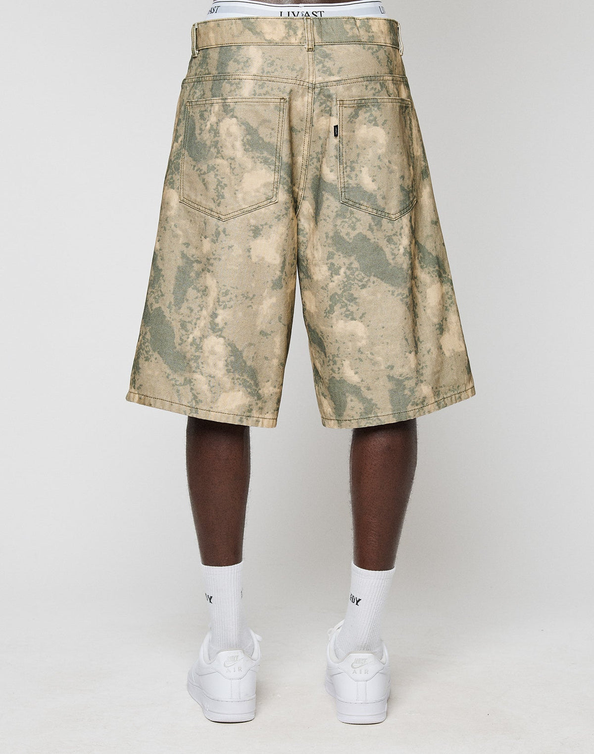A person faces away, wearing LIVE FAST DIE YOUNG Canvas Shorts (100% Baumwolle, Baggy Fit, gerade Bein) with back pockets. The white waistband of underwear shows above the shorts, paired with white Nike socks and sneakers.