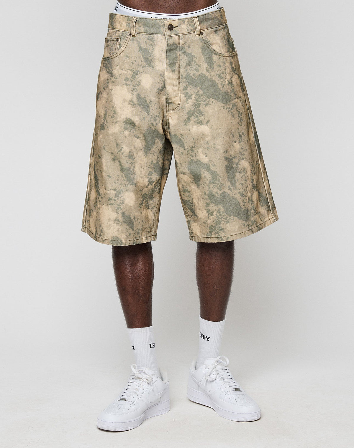 A person stands against a plain light background wearing LIVE FAST DIE YOUNG Canvas Shorts—knee-length, baggy fit, camouflage print, made from 100% Baumwolle—paired with white crew socks and white sneakers.