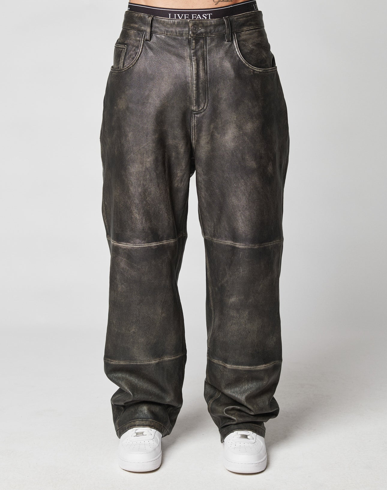 A person wears Baggy Leather Pants by LIVE FAST DIE YOUNG, styled with white sneakers. The pants are dark-washed faux leather with seam details. The LIVE FAST waistband of underwear is visible above the pants. Plain light gray background, flag label shown.
