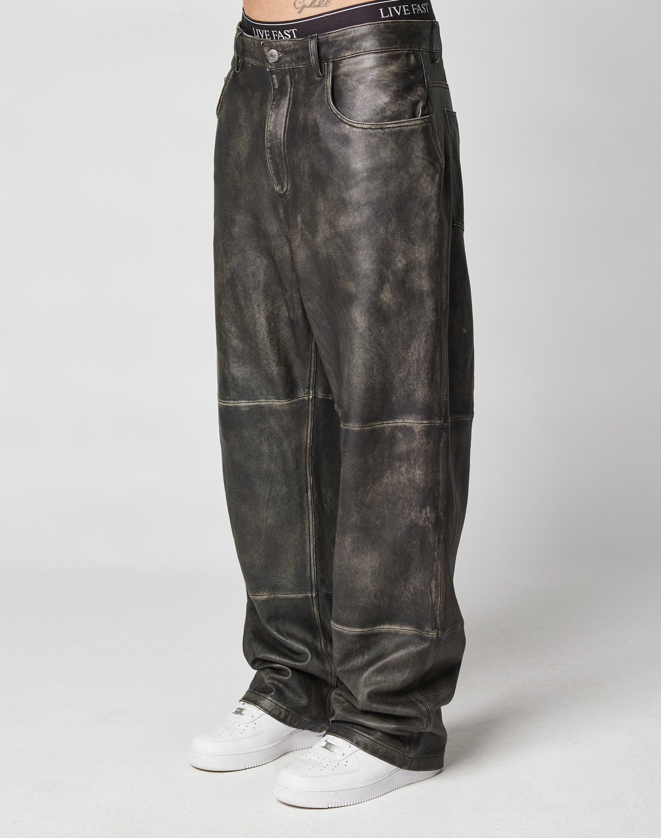 A person showcases the lower body in LIVE FAST DIE YOUNG Baggy Leather Pants—dark distressed, visible seams, and vintage style—paired with white sneakers against a plain background. | model