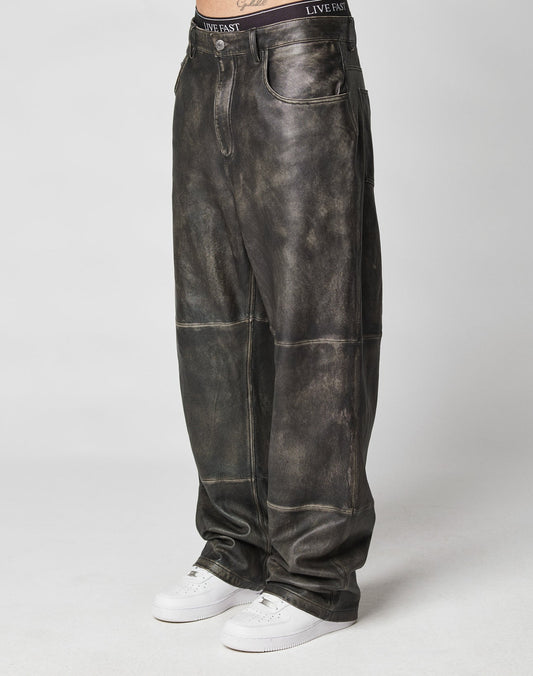 A person showcases the lower body in LIVE FAST DIE YOUNG Baggy Leather Pants—dark distressed, visible seams, and vintage style—paired with white sneakers against a plain background. | model