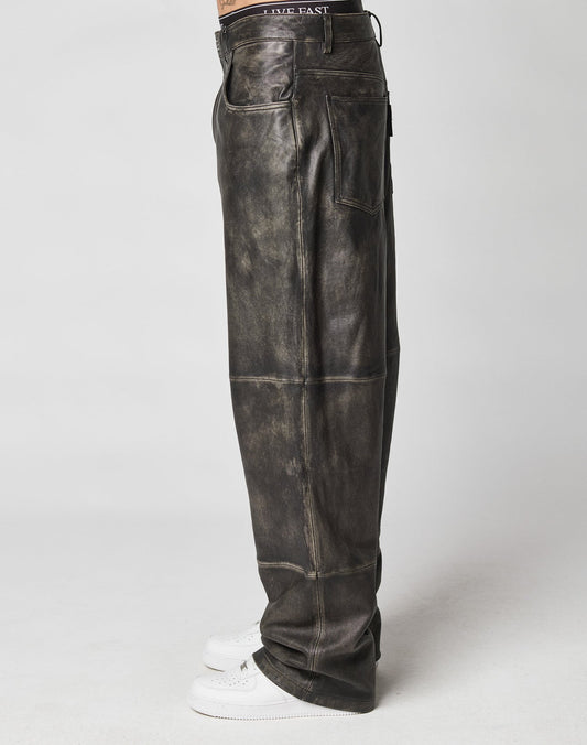 Side view of a person in baggy fit, distressed black Baggy Leather Pants by LIVE FAST DIE YOUNG with a flag label, white sneakers, and a black waistband that features “LIVE FAST,” standing against a plain light background.