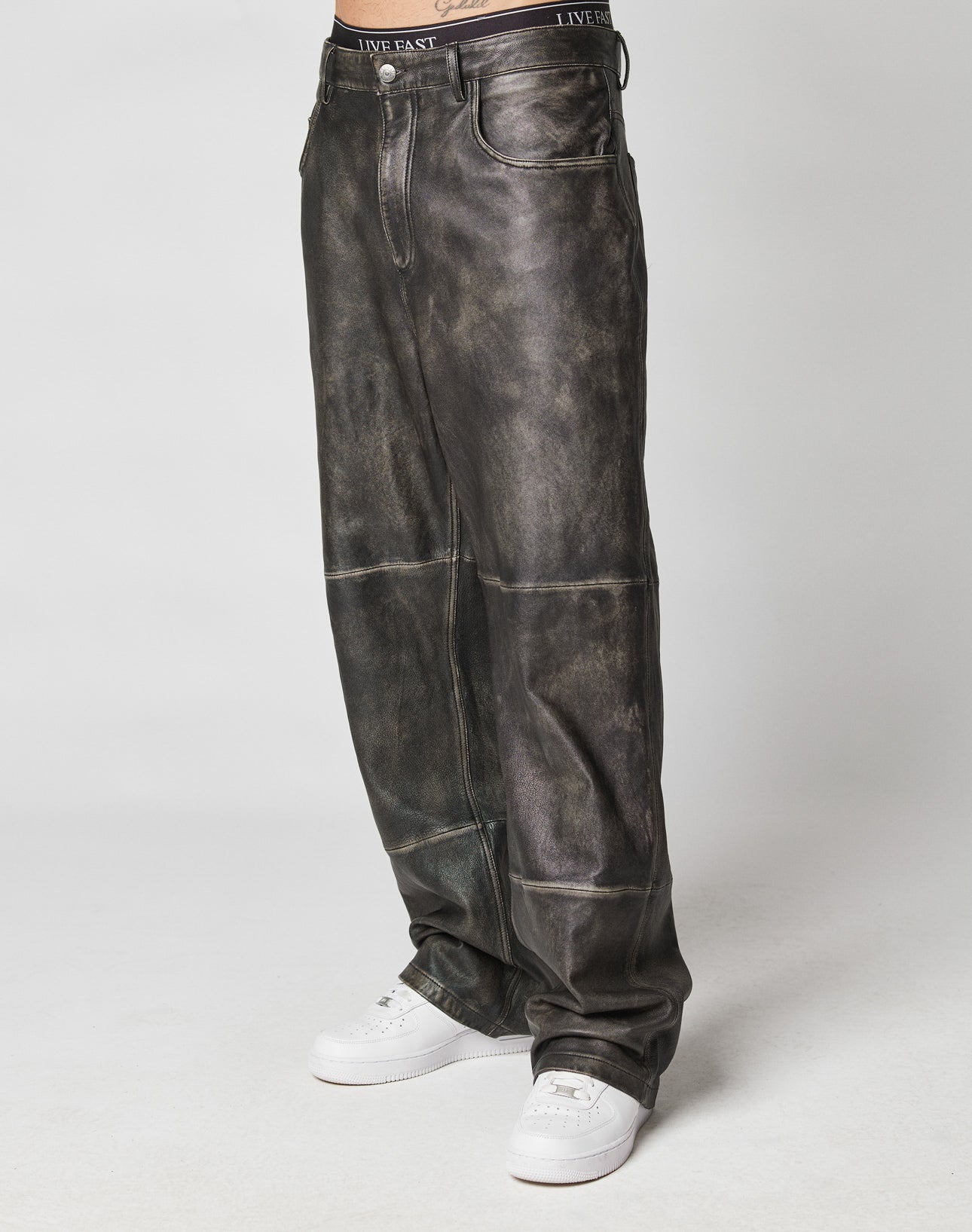 Someone wearing baggy black Baggy Leather Pants by LIVE FAST DIE YOUNG with a Flag Label detail and LIVE FAST waistband, paired with white sneakers, stands against a plain light background.