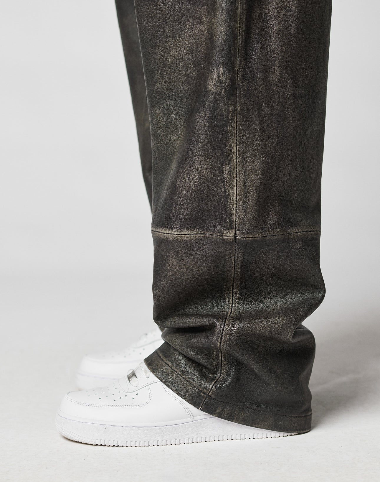 A person wearing LIVE FAST DIE YOUNG Baggy Leather Pants and white sneakers is photographed from the knee down against a plain light background.