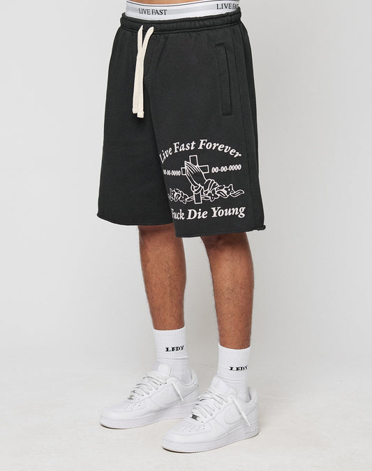 A person wears LFDY Praying Shorts by LIVE FAST DIE YOUNG—regular fit, black with white “Live Fast Forever, Fuck Die Young” text and rose graphic, elastic waistband, side pockets—paired with “LDY” socks and white sneakers. | model