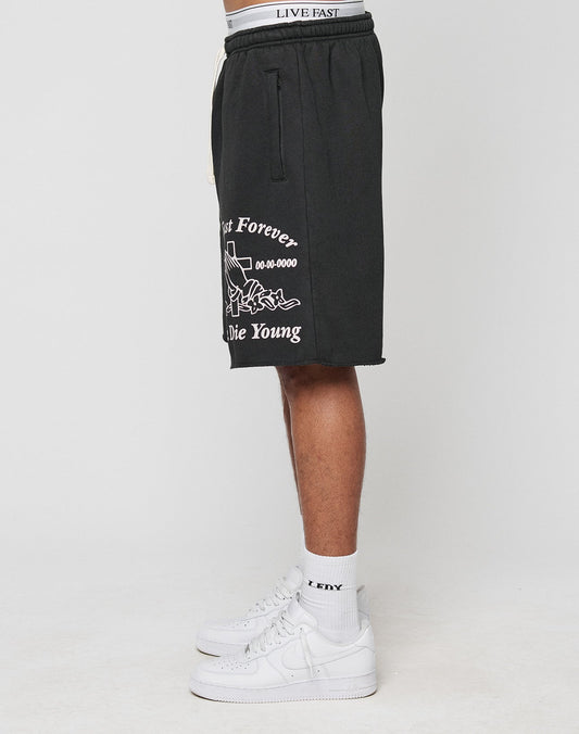 A person stands sideways wearing black LIVE FAST DIE YOUNG LFDY Praying Shorts with elastic waistband and side pockets, plus white LFDY socks and white sneakers. The shorts display a graphic with Last Forever, Die Young. Only the lower body is visible.