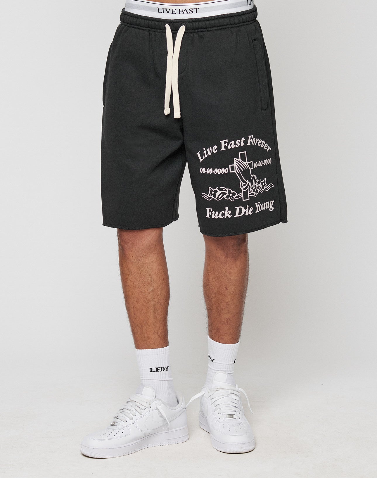 A person wearing LFDY Praying Shorts by LIVE FAST DIE YOUNG with bold text, graphic prints, seitliche Eingrifftaschen, and Elastikbund in regular fit, styled with white socks and sneakers against a plain background.