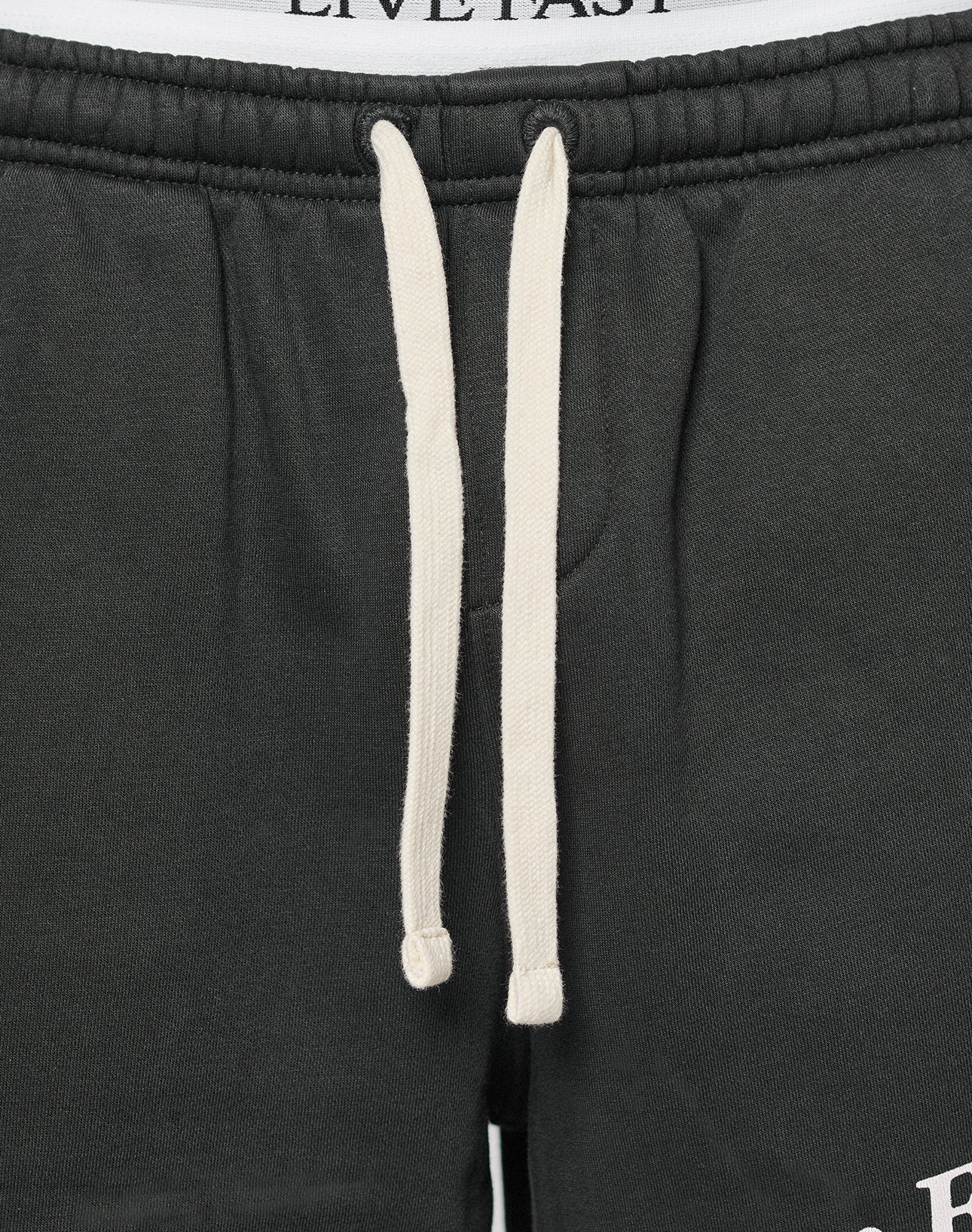 Close-up of the waistband on LFDY Praying Shorts by LIVE FAST DIE YOUNG, featuring black fabric, white drawstrings, an elastic waistband, and convenient side pockets. The shirt above shows part of the LIVE FAST logo.