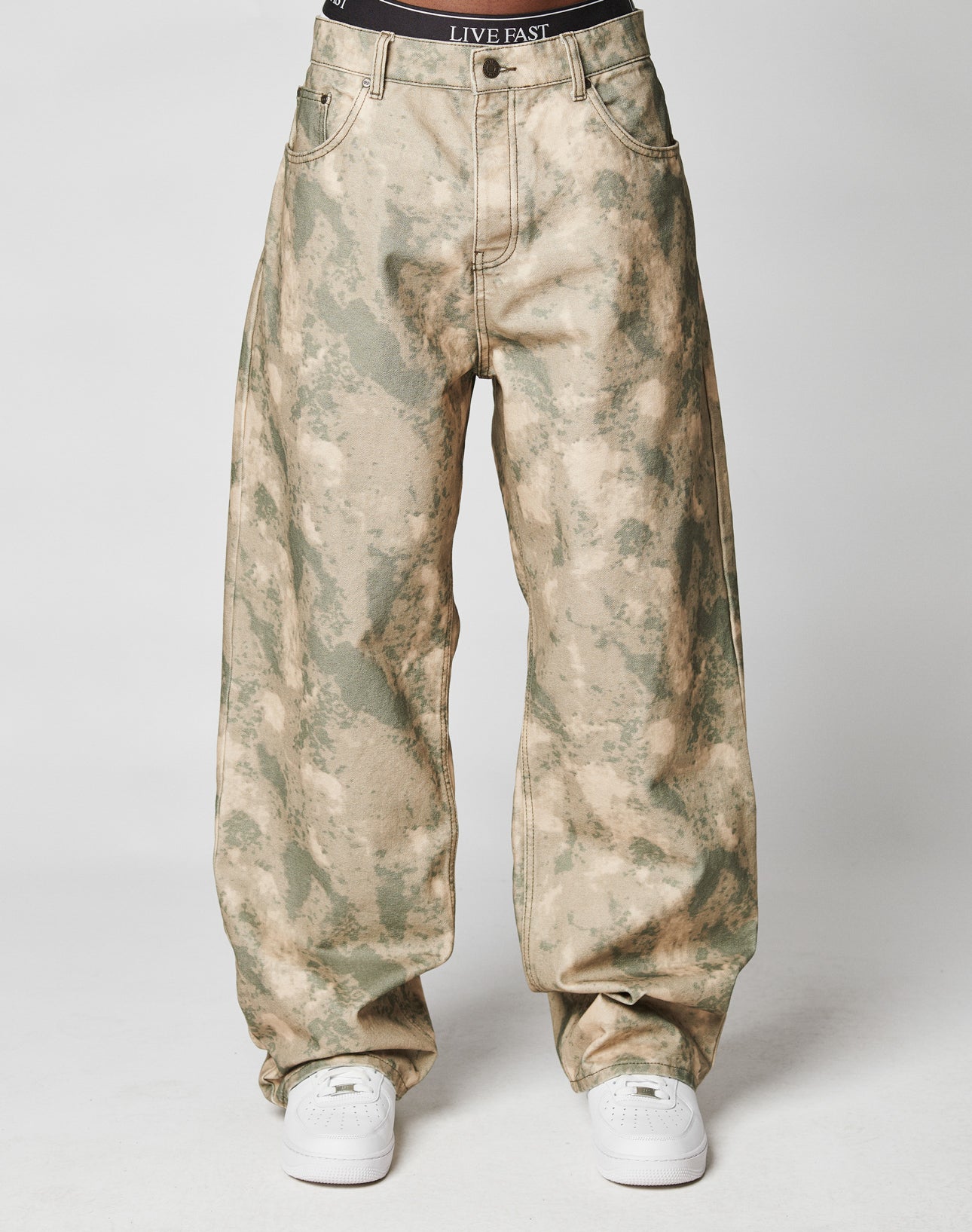 A model wears loose-fitting green and beige camouflage Live Fast Canvas Pants by LIVE FAST DIE YOUNG, paired with white sneakers and black LIVE FAST waistband underwear, posing against a plain white background.