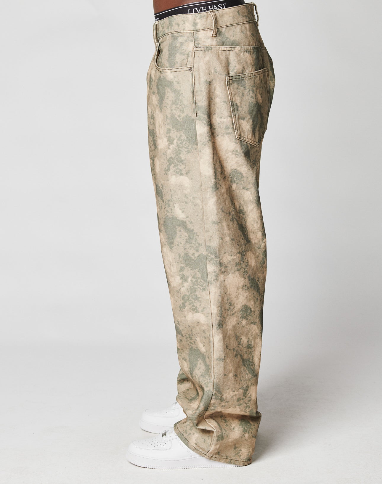 A person in LIVE FAST DIE YOUNG Live Fast Canvas Pants—baggy-fit, 100% cotton camouflage with an elastic waistband and large side pockets—plus white sneakers, standing in profile against a plain white background.