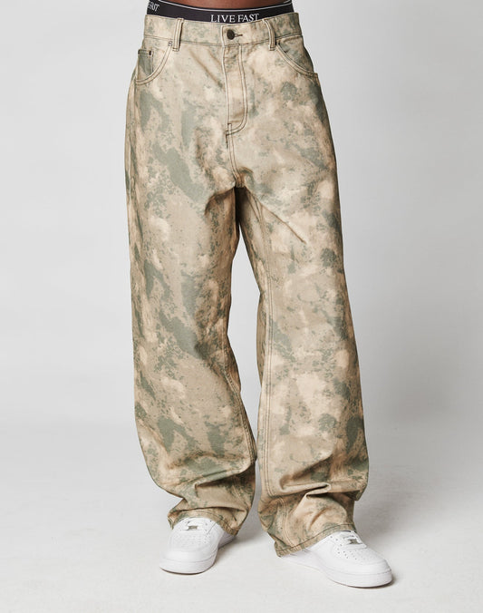 A person stands against a plain white background wearing LIVE FAST DIE YOUNGs Live Fast Canvas Pants in loose-fitting beige and green camouflage, featuring a gerades Bein and Baggy Fit, paired with white sneakers and a black I LIVE FAST waistband.