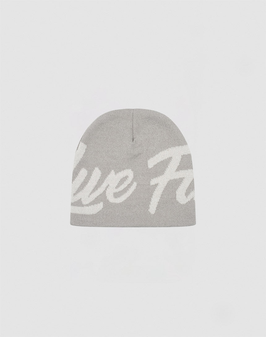 The LFDY Jacquard Beanie by LIVE FAST DIE YOUNG is a light gray knit beanie made from 100% Polyacryl, with large white cursive text partially visible across the front, shown against a plain white background.| plain