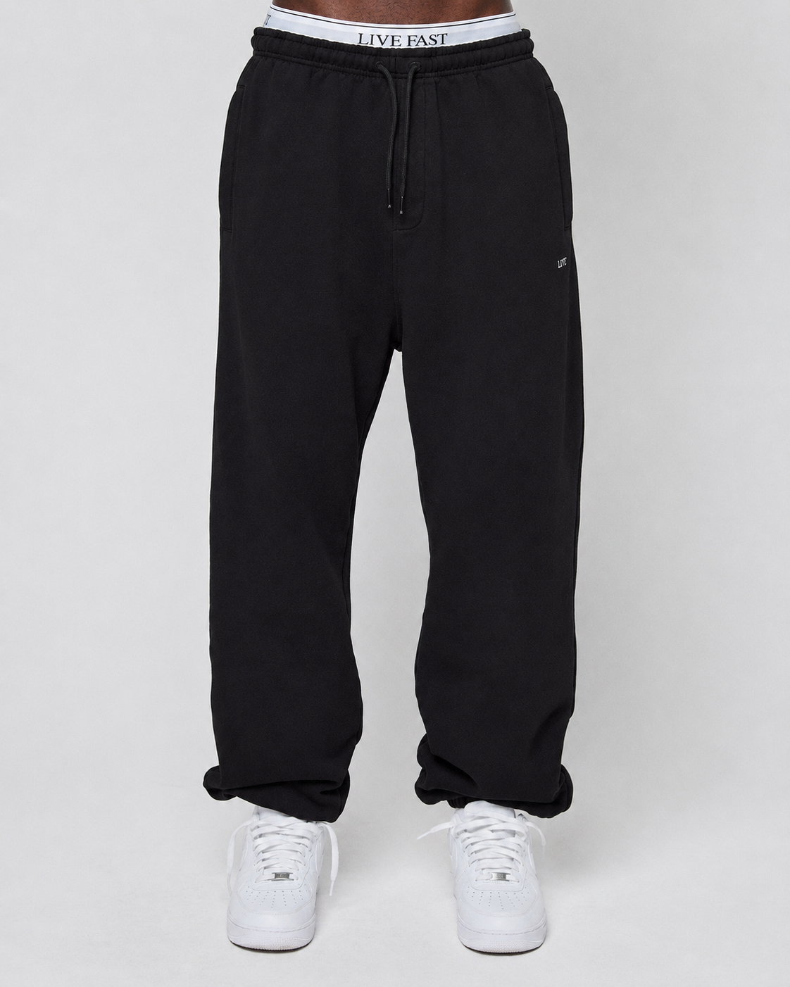 A person in a Basic Loose Jogger by LIVE FAST DIE YOUNG, showing its elastic waistband with a white LIVE FAST logo and paired with white sneakers, stands against a plain light gray background.