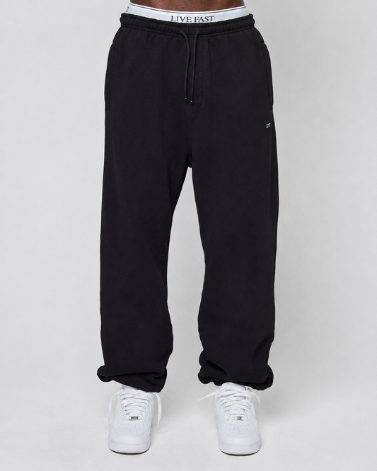 A person in a Basic Loose Jogger by LIVE FAST DIE YOUNG, showing its elastic waistband with a white LIVE FAST logo and paired with white sneakers, stands against a plain light gray background.