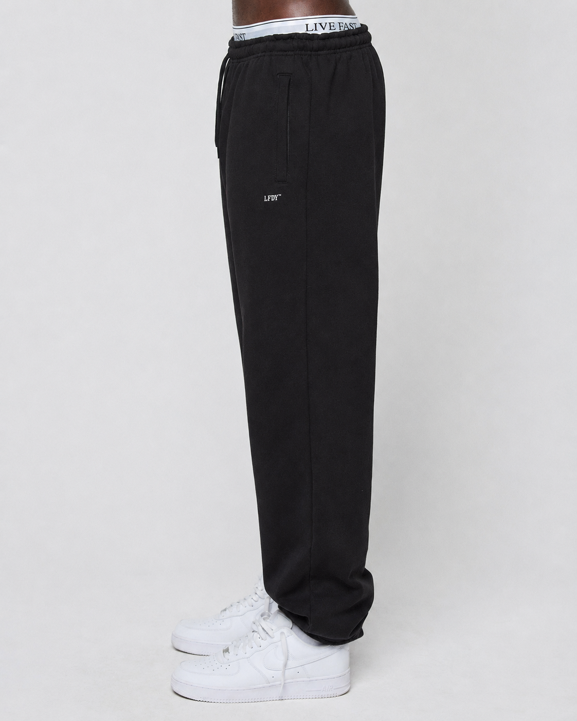 A person stands sideways in black Basic Loose Jogger from LIVE FAST DIE YOUNG, featuring a logo print and elastic waistband, paired with white sneakers against a plain light background.