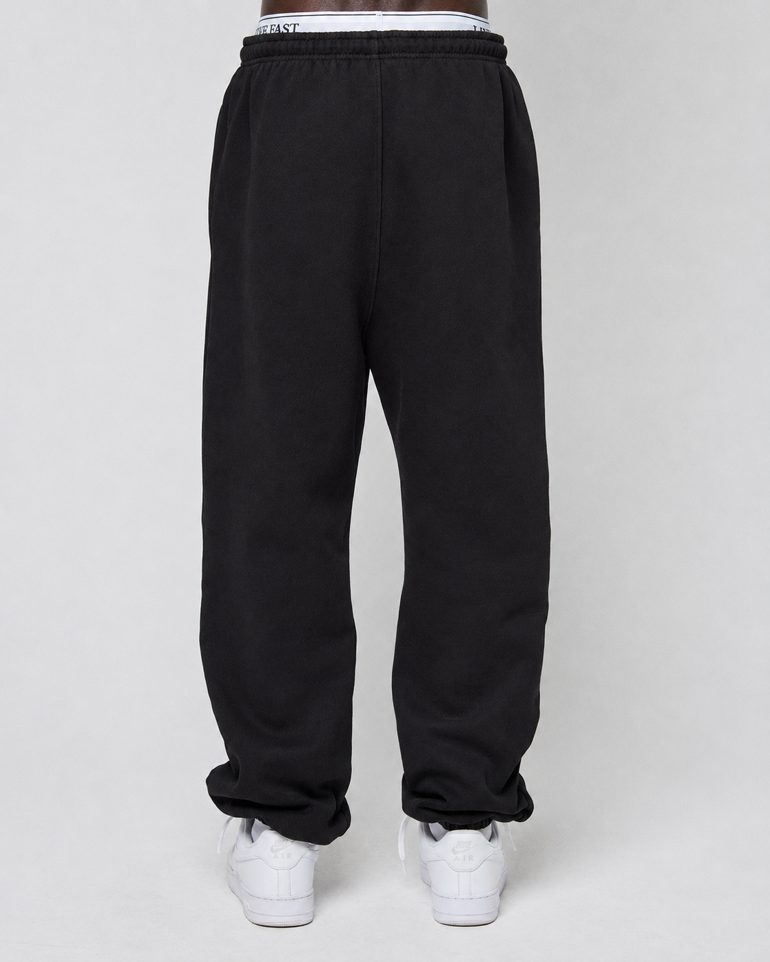The lower half of a person wearing LIVE FAST DIE YOUNG Basic Loose Jogger with elastic waistband and white sneakers is shown against a plain light gray background.