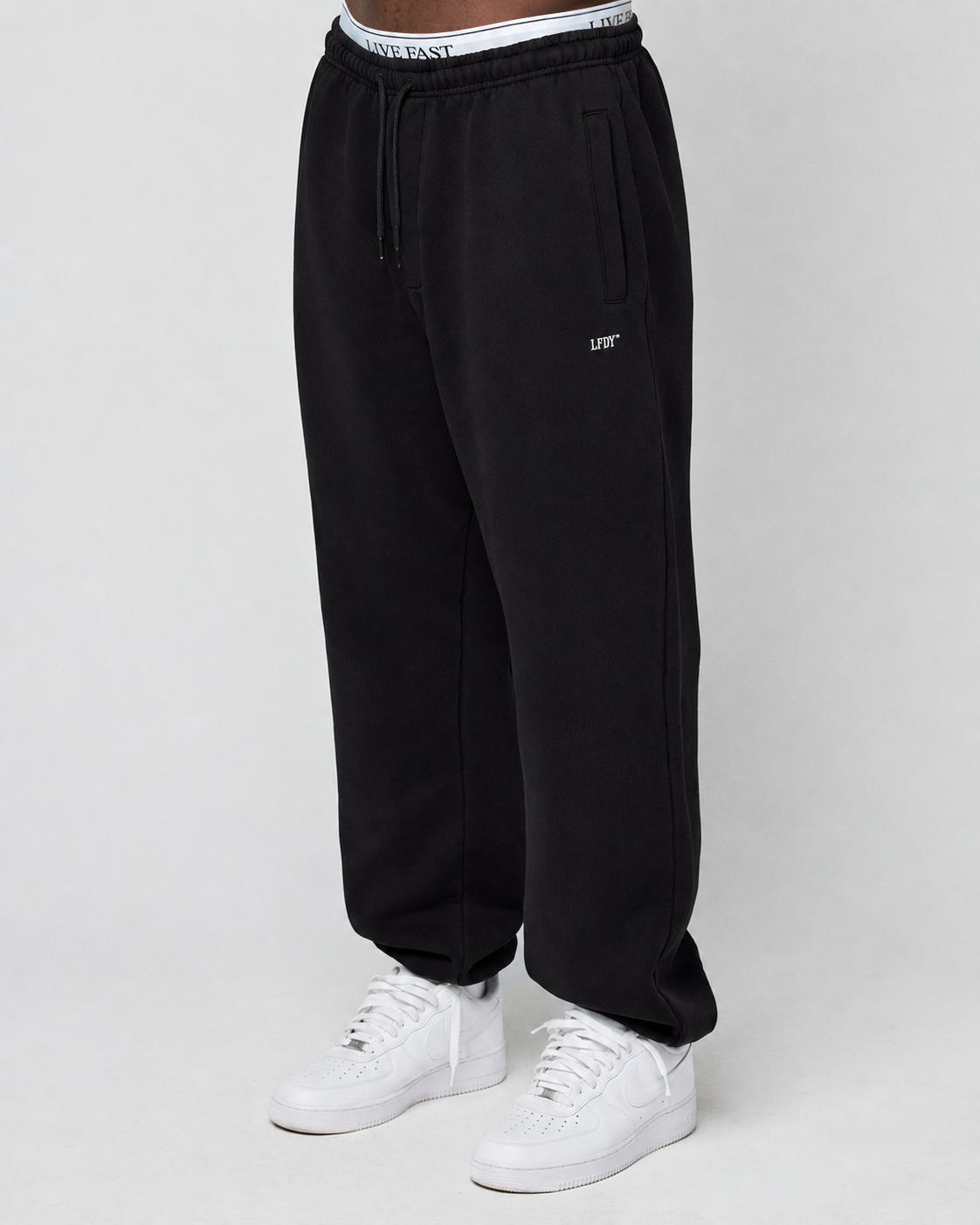 A person wears a Basic Loose Jogger by LIVE FAST DIE YOUNG with “LEFT” logo on the left leg, white high-top sneakers, and an elastic waistband featuring “LIVE FAST.” The background is plain and light-colored. | model