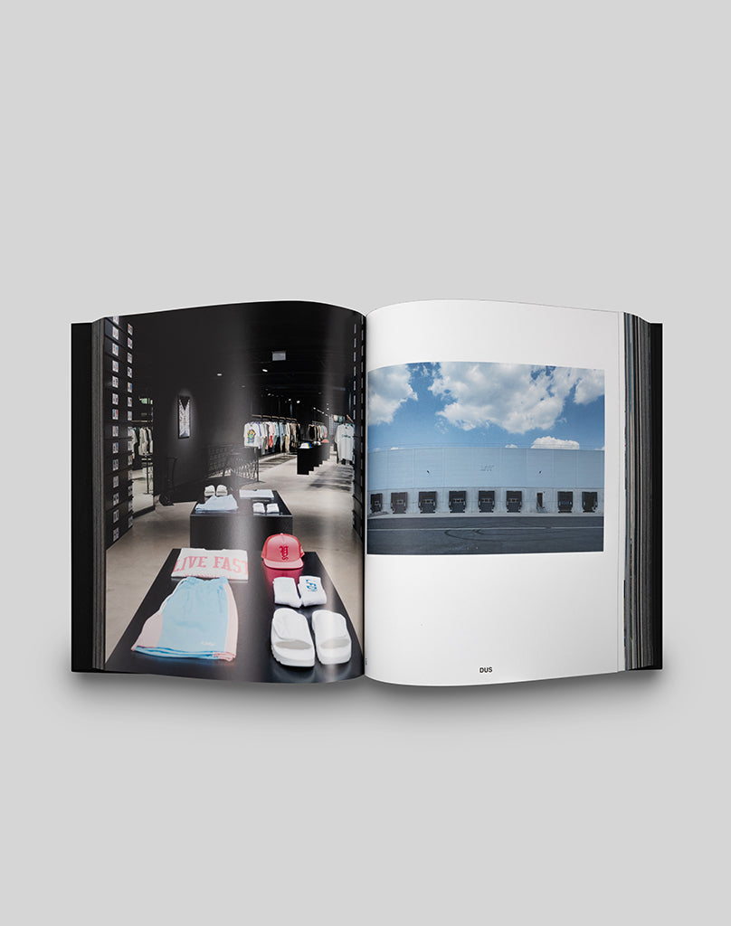 The LFDY Brand Bible by LIVE FAST DIE YOUNG is an open hardcover book showing folded clothes and accessories on the left page, and a white heritage building under a blue sky with clouds on the right—like a visual brand manifesto.