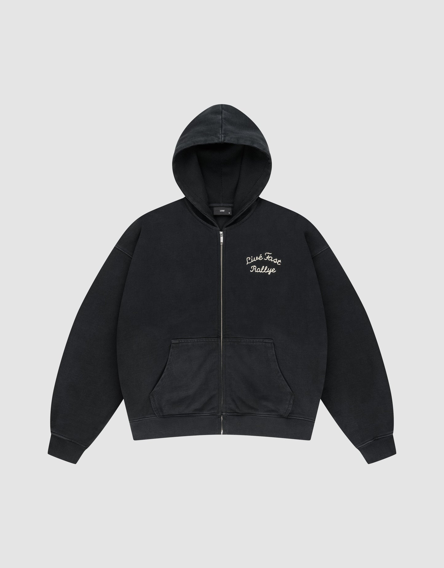 The LFDY L’Équipe Hooded Zip by LIVE FAST DIE YOUNG is a black oversized hoodie with a front pocket, hood, cozy waffle innenfutter, and white embroidered "Pink Face Rallye" text on the left chest. Laid flat on light gray background.