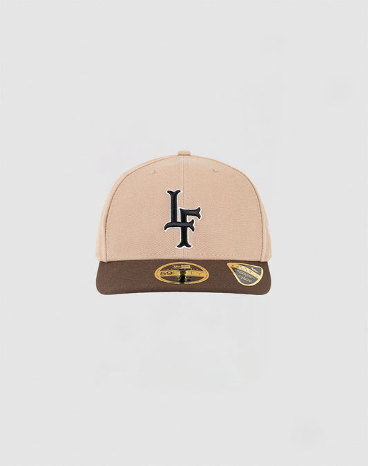 The LFDY x NEW ERA 59FIFTY Low Profile Fitted Cap by LIVE FAST DIE YOUNG features a beige and brown design, a black LF logo on the front, and two gold-and-black stickers on the brown brim, displayed against a white background. 