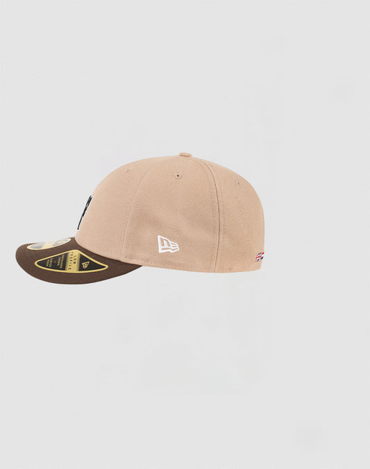 The LFDY x NEW ERA 59FIFTY Low Profile by LIVE FAST DIE YOUNG is shown from the side in tan and brown, featuring a white embroidered New Era logo, small flag detail near the back, and gold brim stickers on a plain light gray background.