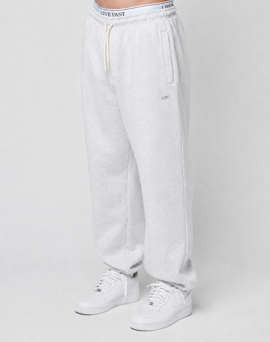 A person wears the LIVE FAST DIE YOUNG Basic Loose Jogger in light gray, featuring a logo print waistband with white drawstring, side pockets, and small embroidered detail on the left thigh, paired with white sneakers against a plain background. | model