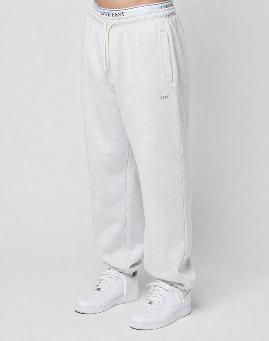 A person wears the LIVE FAST DIE YOUNG Basic Loose Jogger in light gray, featuring a logo print waistband with white drawstring, side pockets, and small embroidered detail on the left thigh, paired with white sneakers against a plain background. | model