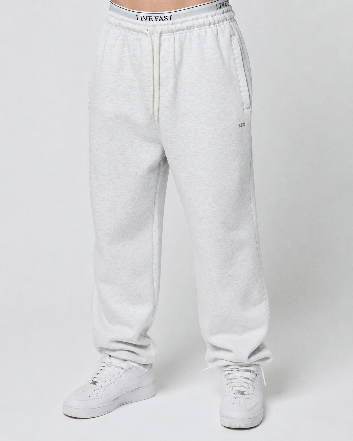 A person in light gray Basic Loose Jogger sweatpants by LIVE FAST DIE YOUNG, featuring an Elastik-Bund waist with a bold LIVE FAST logo, stands against a plain light background paired with white sneakers.