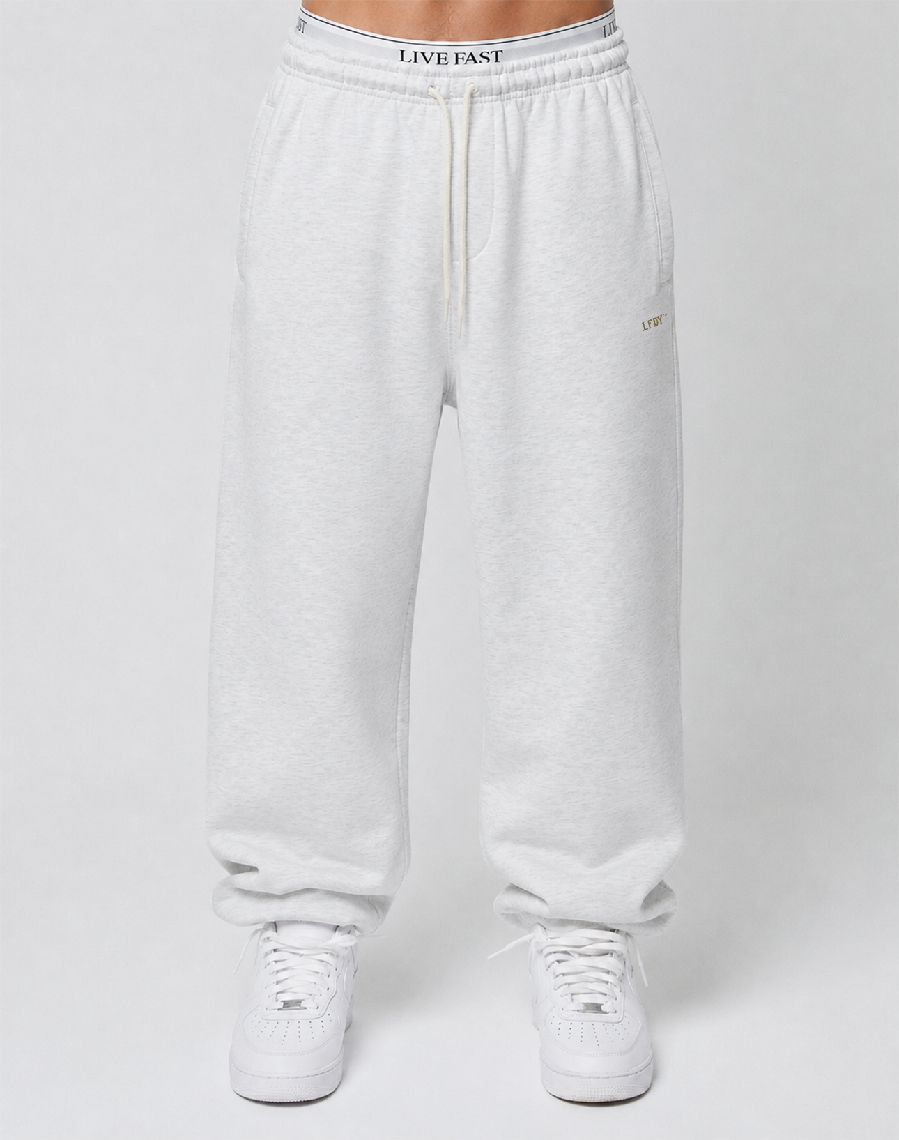 A person wears Basic Loose Jogger sweatpants by LIVE FAST DIE YOUNG with an elastic waistband and logo print, paired with white sneakers, standing against a plain light gray background. Only the lower body is visible.