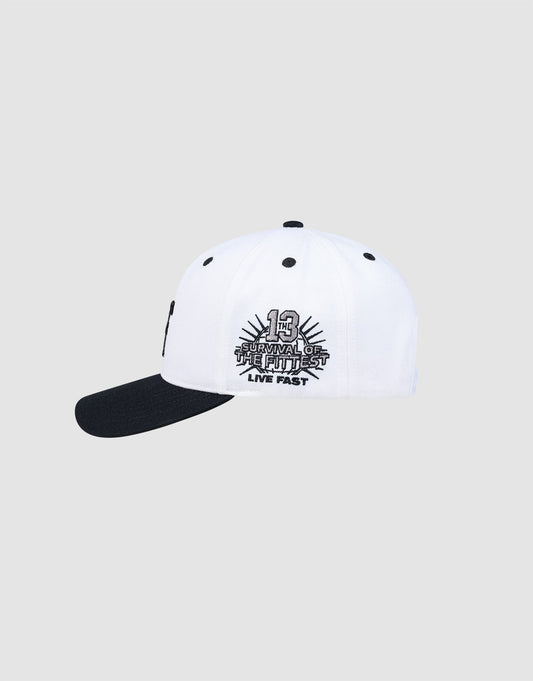 The LFDY Snapback by LIVE FAST DIE YOUNG is a white baseball cap with a black brim and green underside. Features embroidered “13 Survival of the Fittest LIVE FAST” with sunburst on the side and an adjustable snap closure. Plain light gray background.