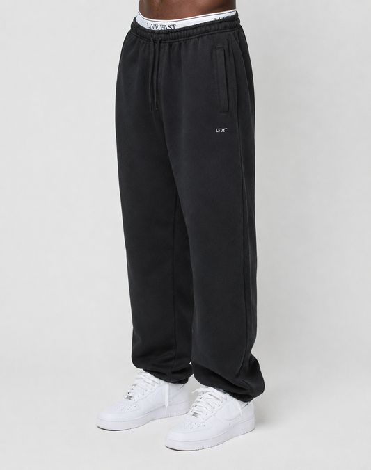 A person is wearing the Basic Loose Jogger by LIVE FAST DIE YOUNG, featuring black fabric, an Elastik-Bund drawstring waist, a visible white LIVE FAST waistband, and white sneakers, standing against a plain light background. | model