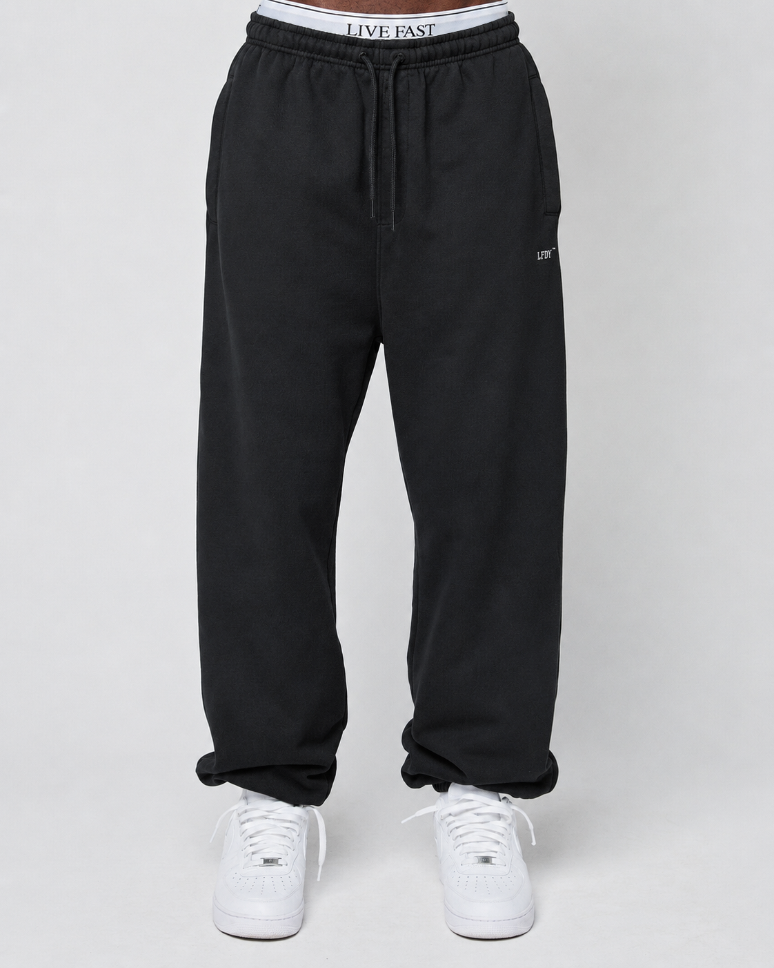 A person wears the Basic Loose Jogger by LIVE FAST DIE YOUNG—black, loose-fit sweatpants with an elastic waistband featuring “LIVE FAST” text, paired with white sneakers, against a plain light background.
